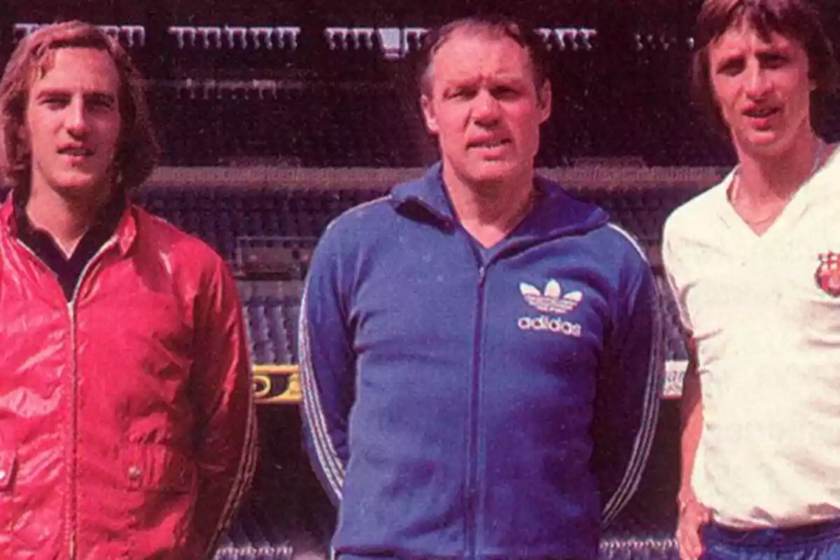 Three men pose together in a football stadium, one wearing a red jacket, another a blue tracksuit and the third a white T-shirt.