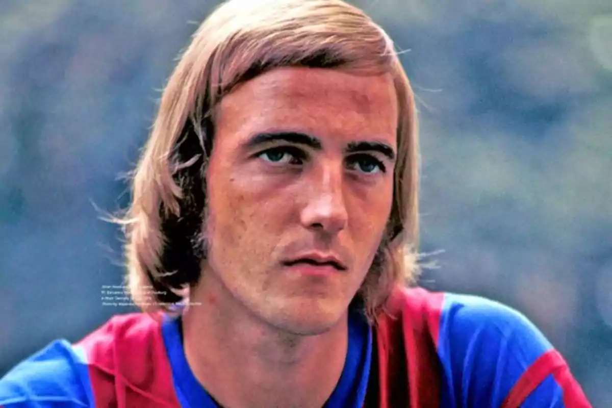 A man with blond hair and a serious expression wears a football shirt with blue and red colors.