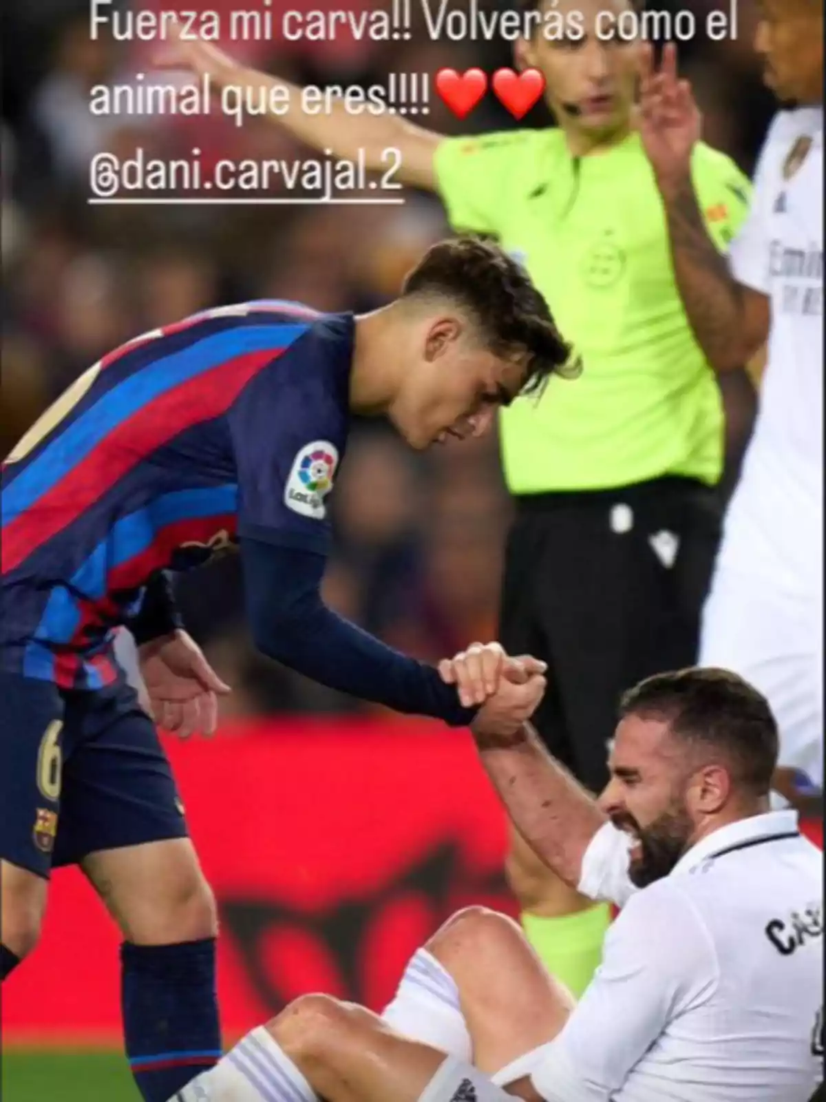 A soccer player helps another to get up during a match, while a referee watches the scene.