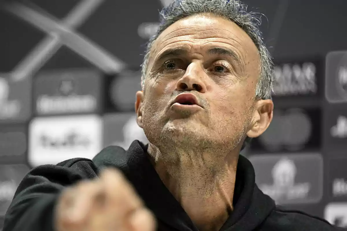 Luis Enrique speaking at a press conference with a background of blurred logos.