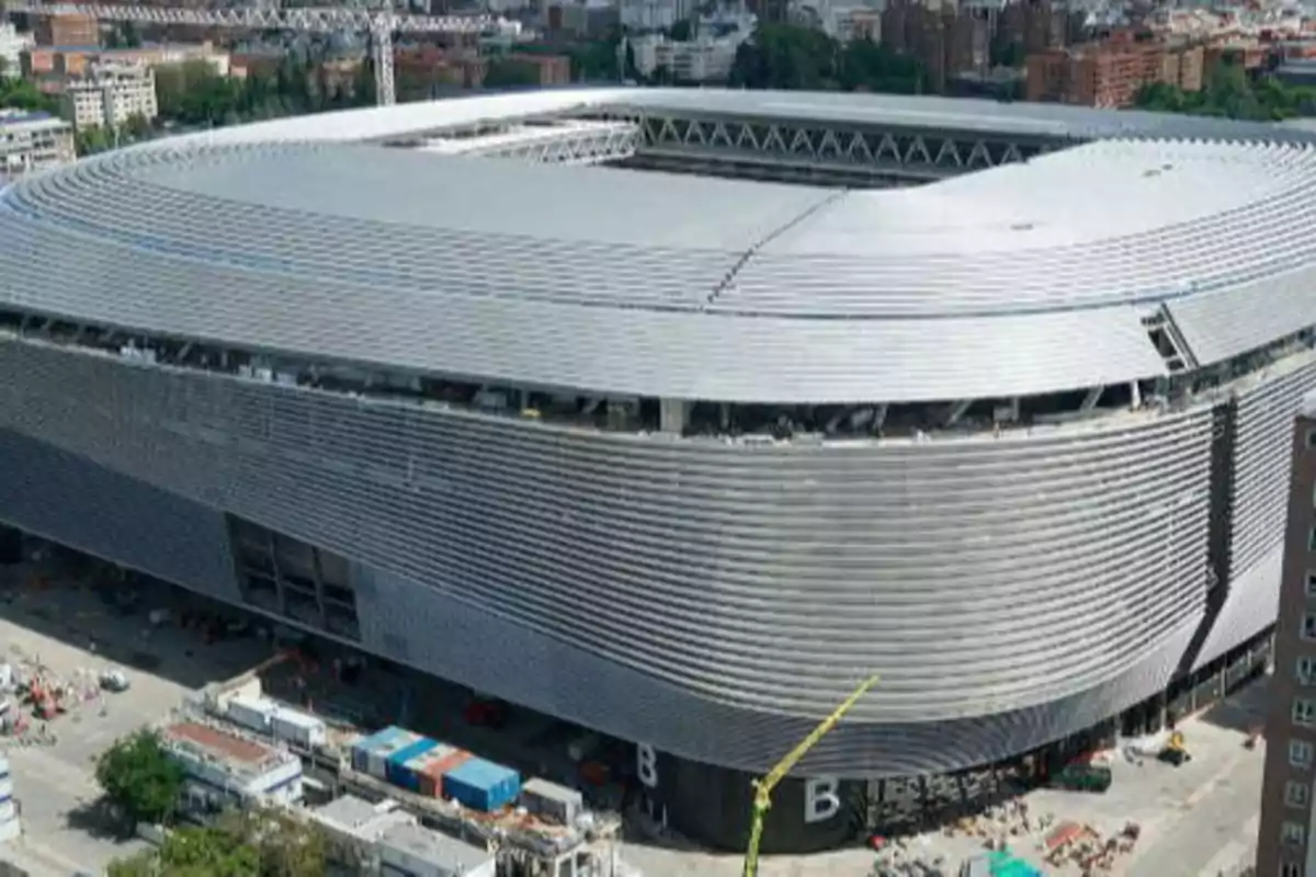 Aerial view of a modern stadium under construction with a corrugated metal structure and an urban environment.