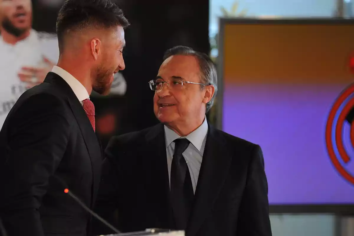 Two men dressed in suits smile as they look at each other at a formal event.