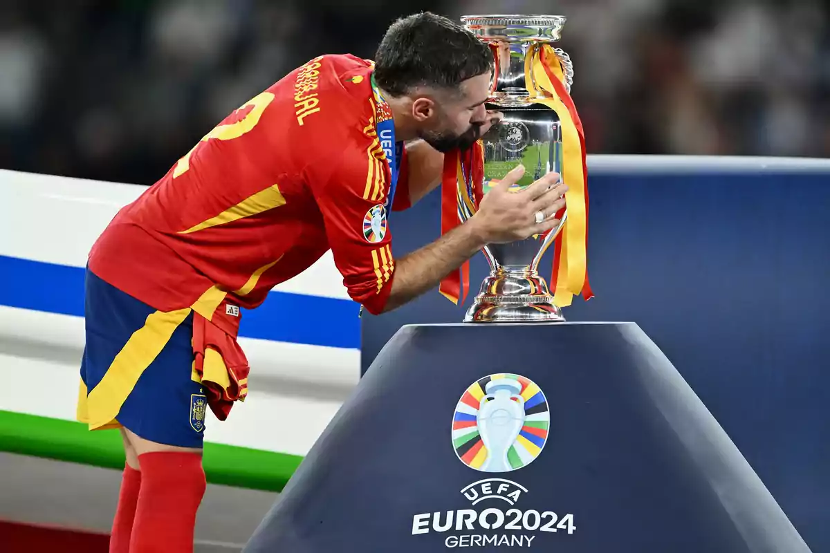 A football player in a red uniform kisses a UEFA Euro 2024 trophy decorated with ribbons.